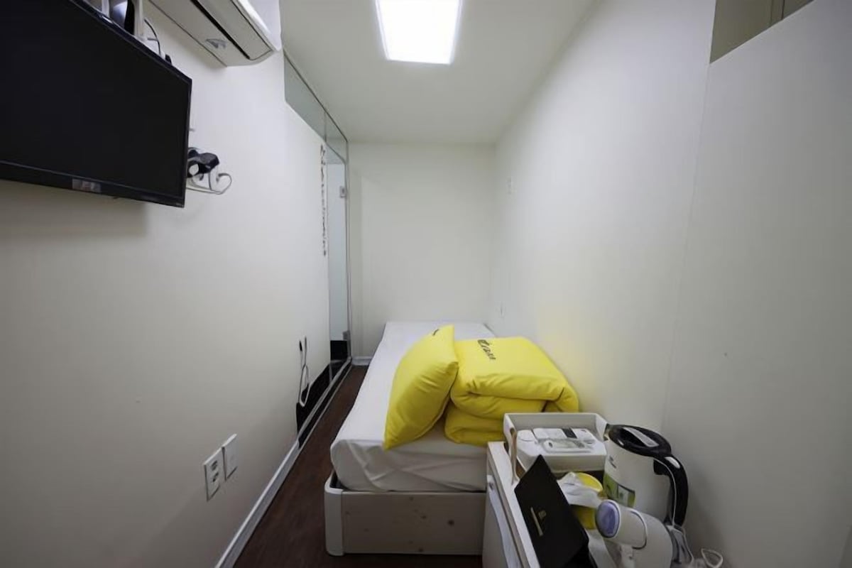 A small, narrow hotel room with a single bed, a bedside table with a lamp, and a wall-mounted TV.