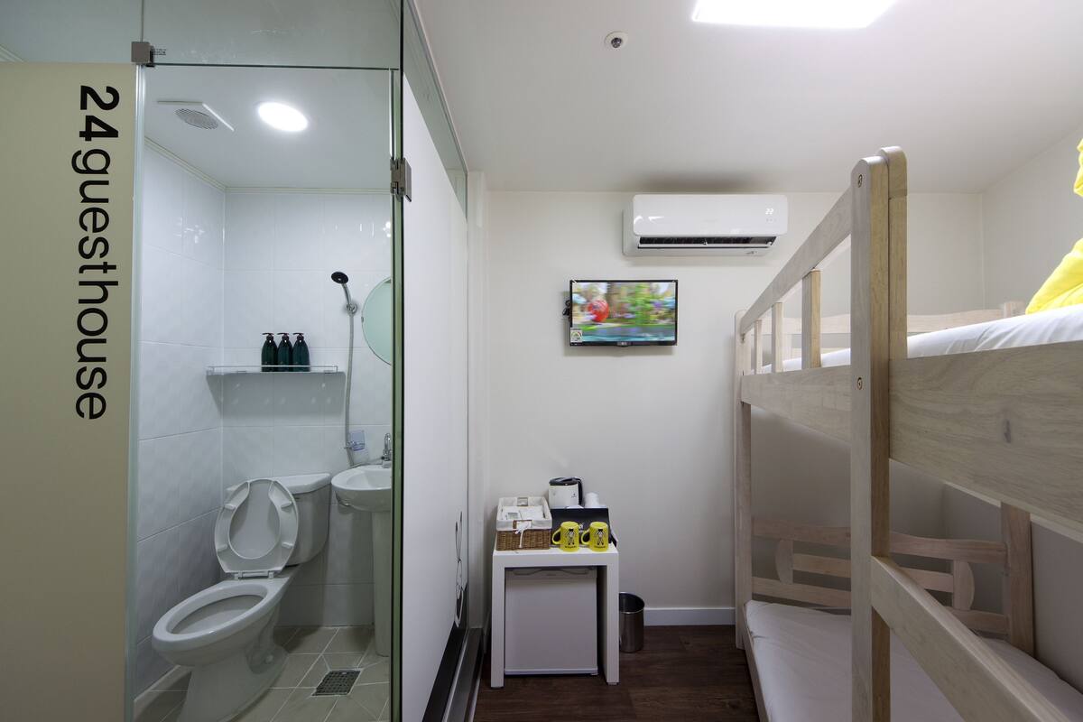 A compact hotel room with a shower, toilet, bunk bed, and a small table with toiletries.