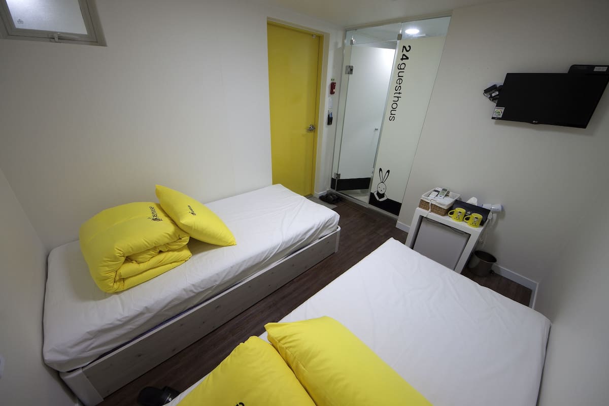 A room with two beds, each with yellow pillows, a small table with a mirror, and a door labeled "24 guests".