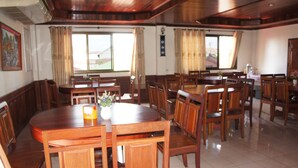 Un comedor amplio con varias mesas y sillas de madera, ventanas grandes con cortinas y un reloj en la pared.