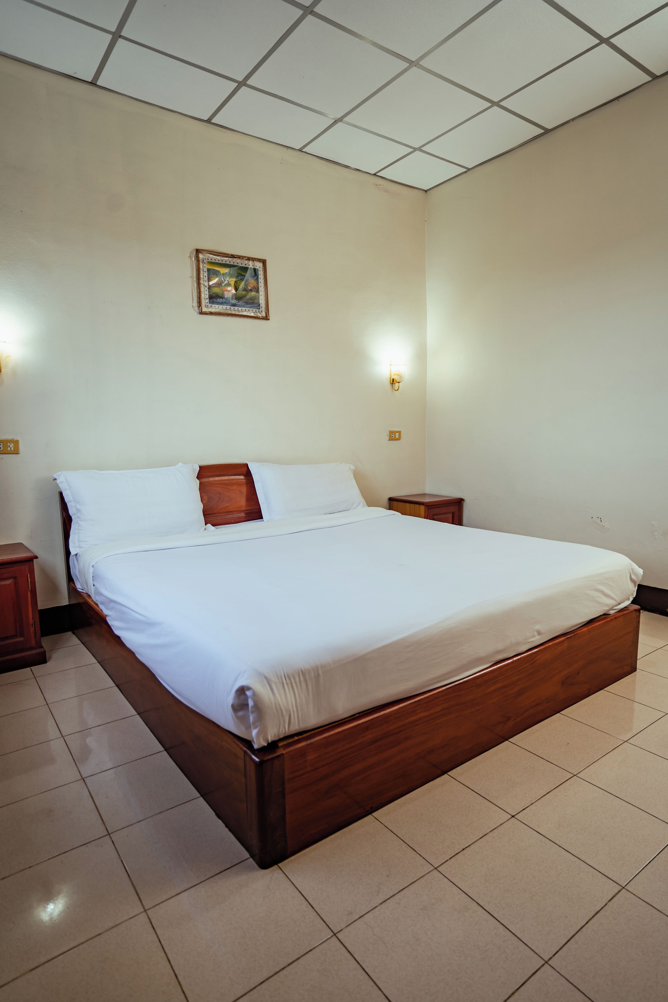 Habitación estándar de hotel con una cama individual, dos mesitas de noche, lámpara de pared y luz de techo.