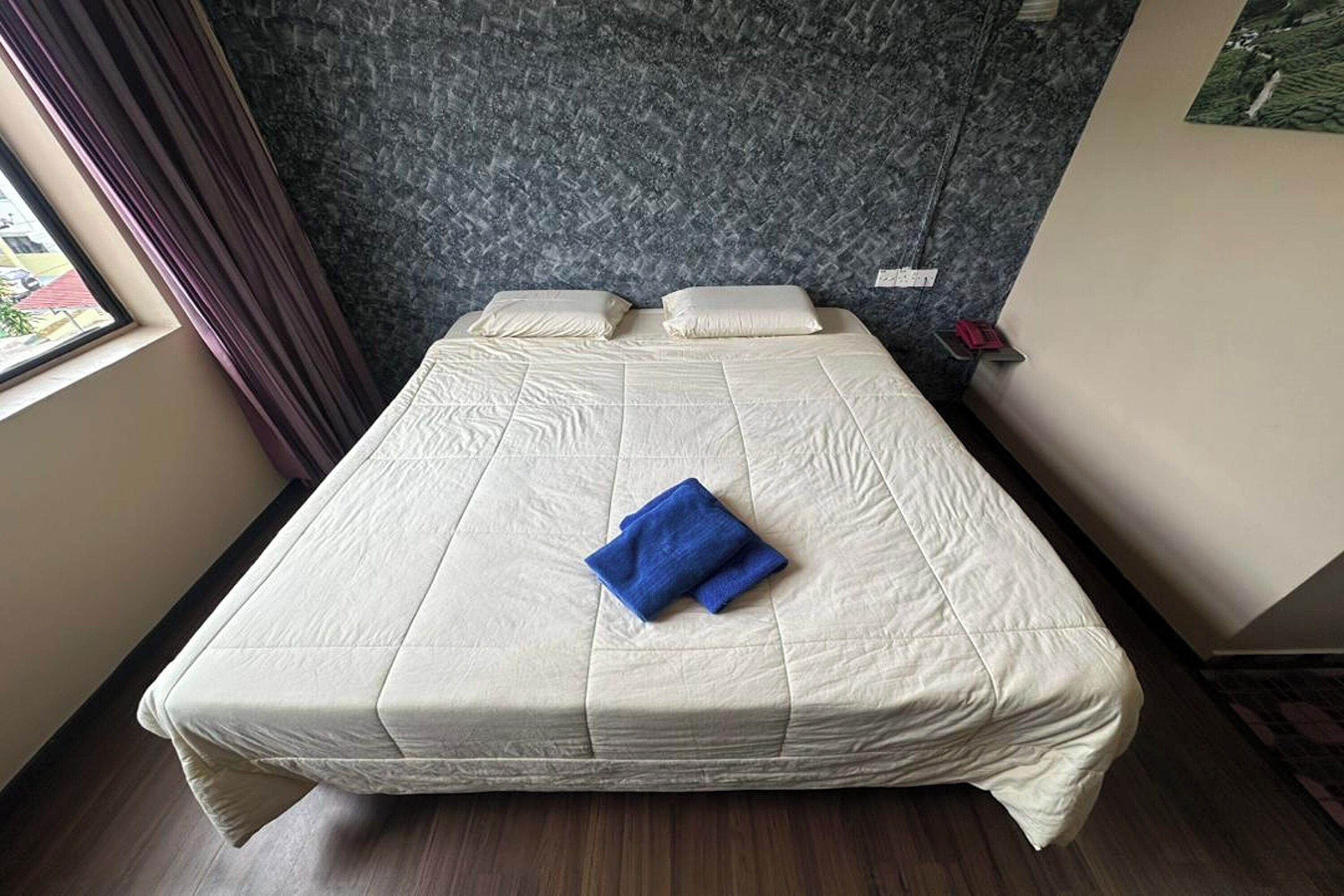 Een netjes opgemaakt bed met een witte dekbedovertrek en twee kussens, een blauwe handdoek op het bed, en een raam met gordijnen aan de linkerzijde.