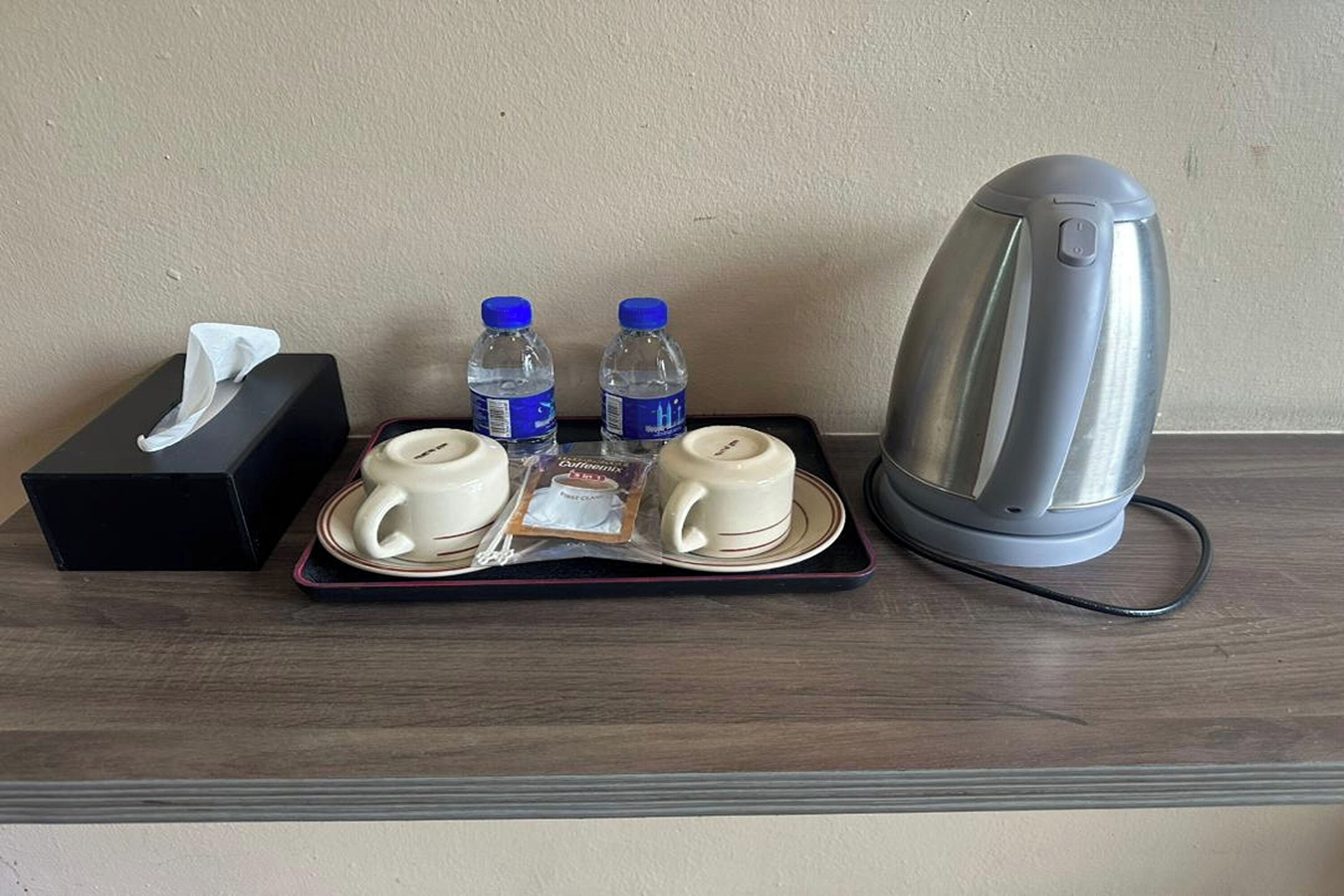 Un plateau contenant deux tasses, deux bouteilles d’eau, une boîte de mouchoirs et une bouilloire, le tout posé sur une surface en bois.