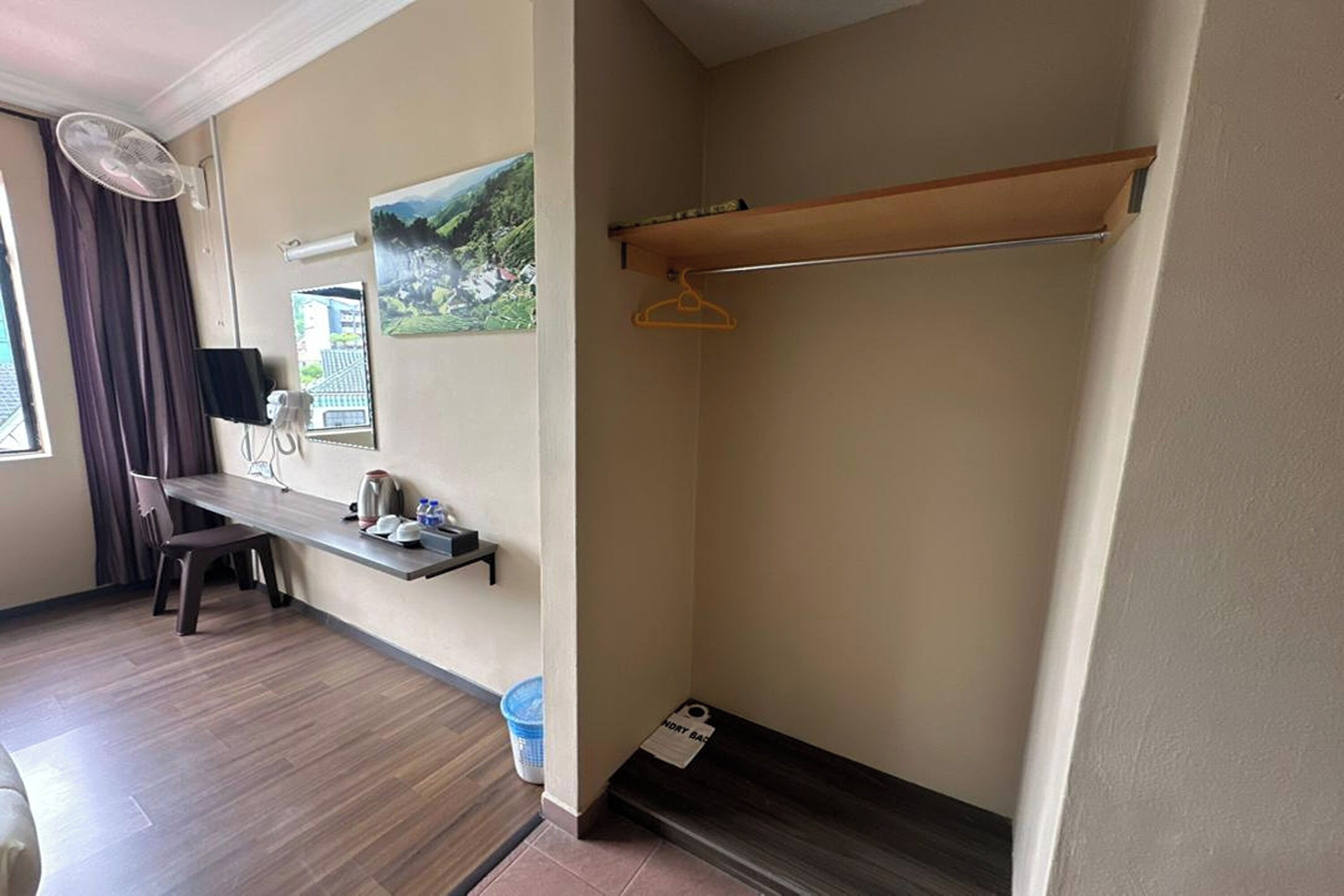 A room with a desk, a hanging coat rack, a window with a view of a landscape, and a fan.