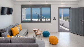 Een moderne woonkamer met een grijze bank, gele en blauwe kussens, een flatscreen-tv en een groot raam met uitzicht op zee.
