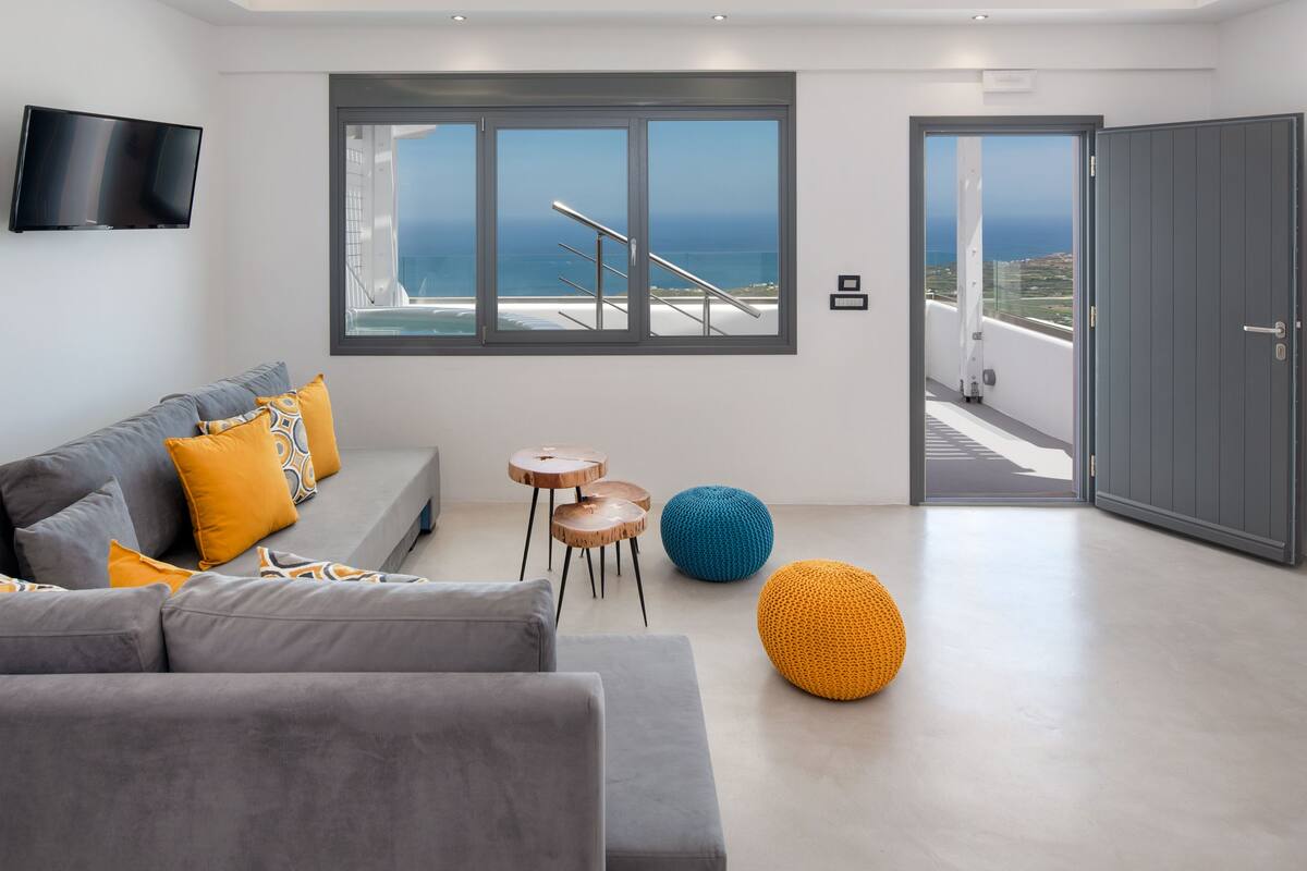 Un salon moderne avec un canapé gris, des coussins jaunes et bleus, un téléviseur à écran plat et une grande fenêtre donnant sur la mer.