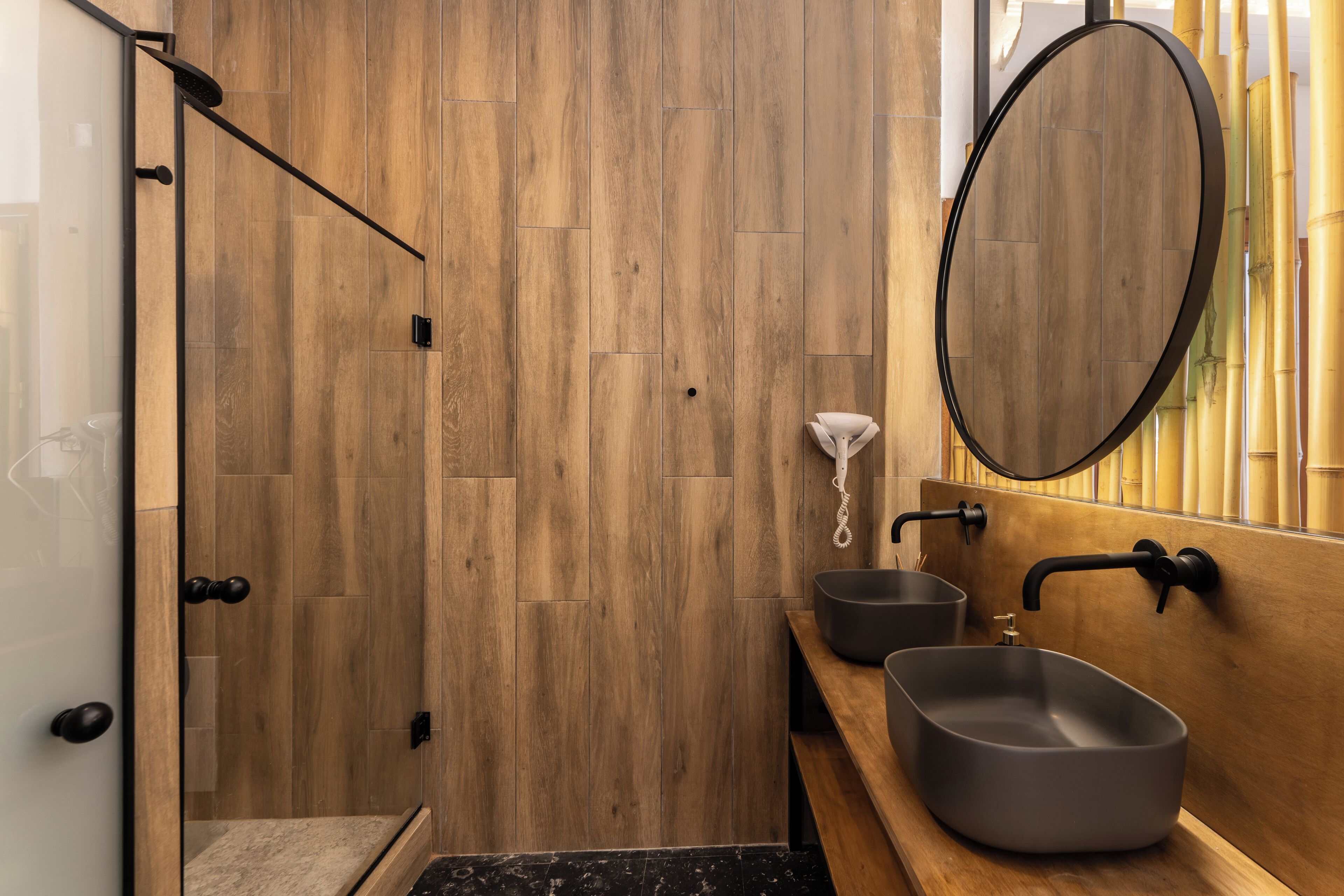 Un baño moderno con paneles de madera, cabina de ducha de vidrio y dos lavamanos con grifería negra.
