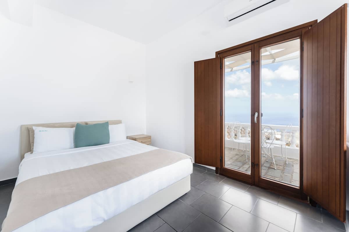 Une chambre à coucher comprenant un lit, une table de chevet, une armoire en bois et une porte-fenêtre coulissante donnant sur un balcon aménagé avec du mobilier d’extérieur.
