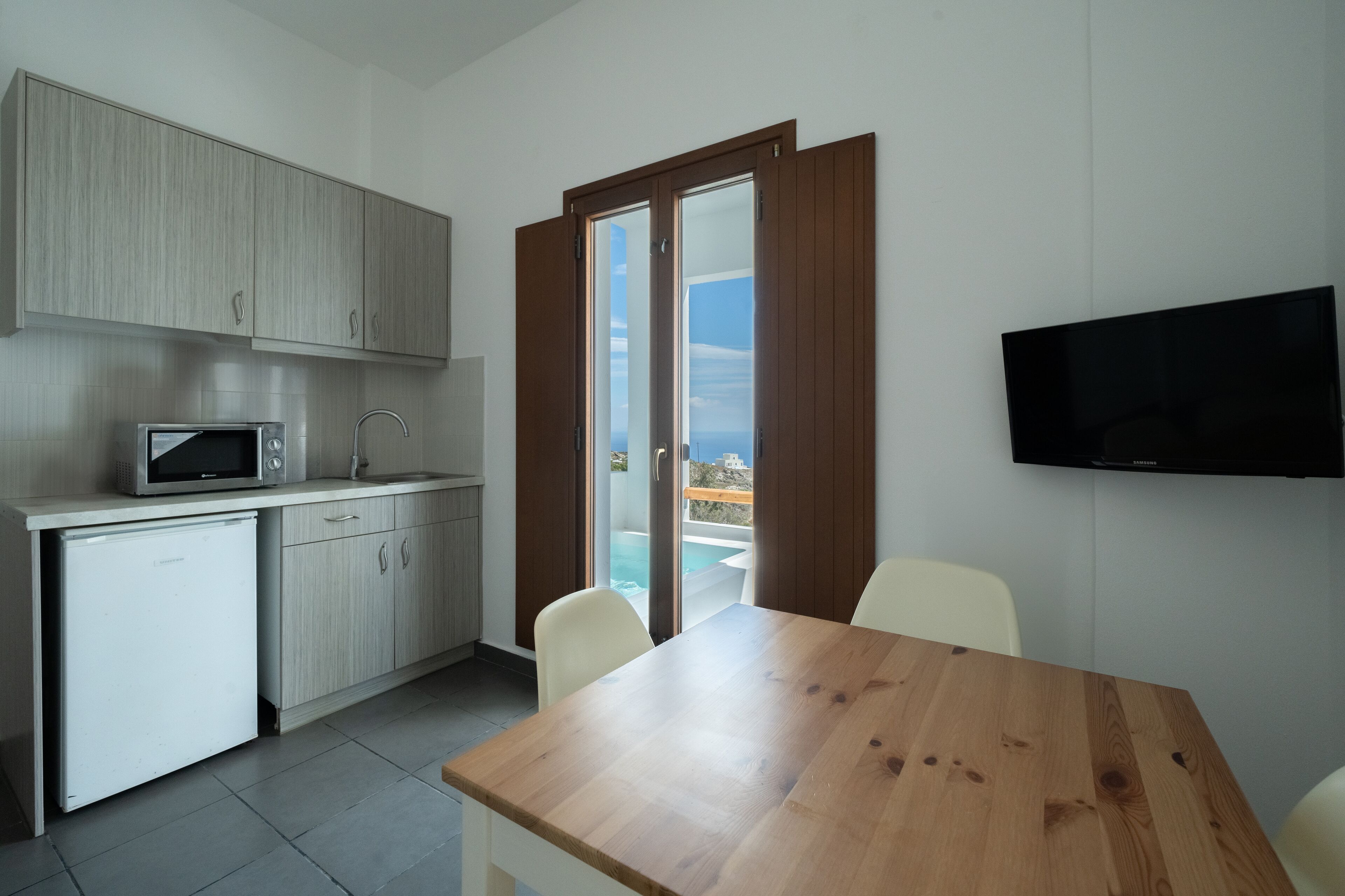 Una cocina funcional con microondas, un refrigerador pequeño y una mesa de comedor con sillas. Un televisor está montado en la pared, y una puerta corrediza de vidrio da a un área exterior con piscina.
