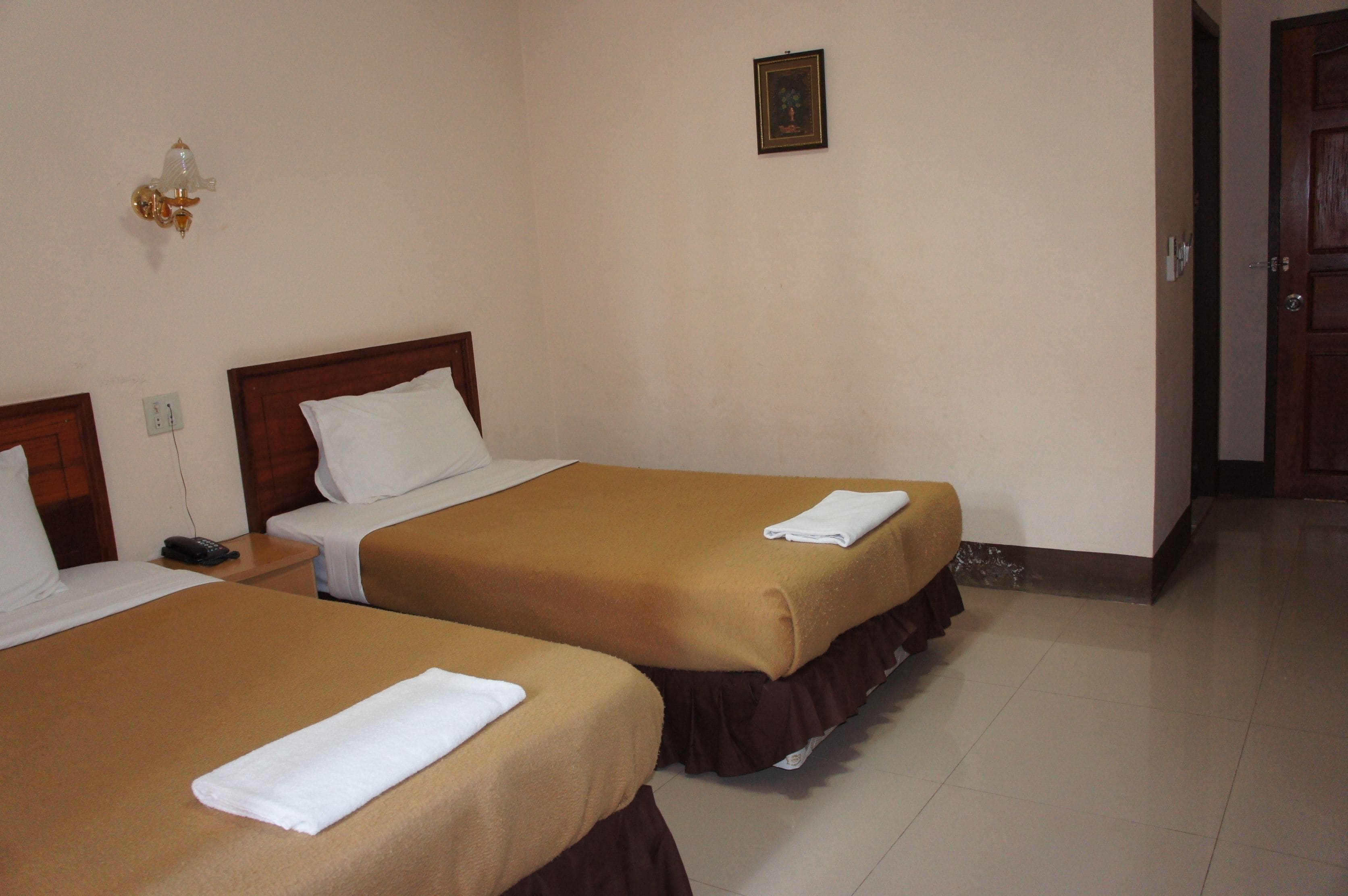 A hotel room with two beds, each with a brown bedspread and white pillows, a bedside table with a phone, and a wall-mounted light fixture.