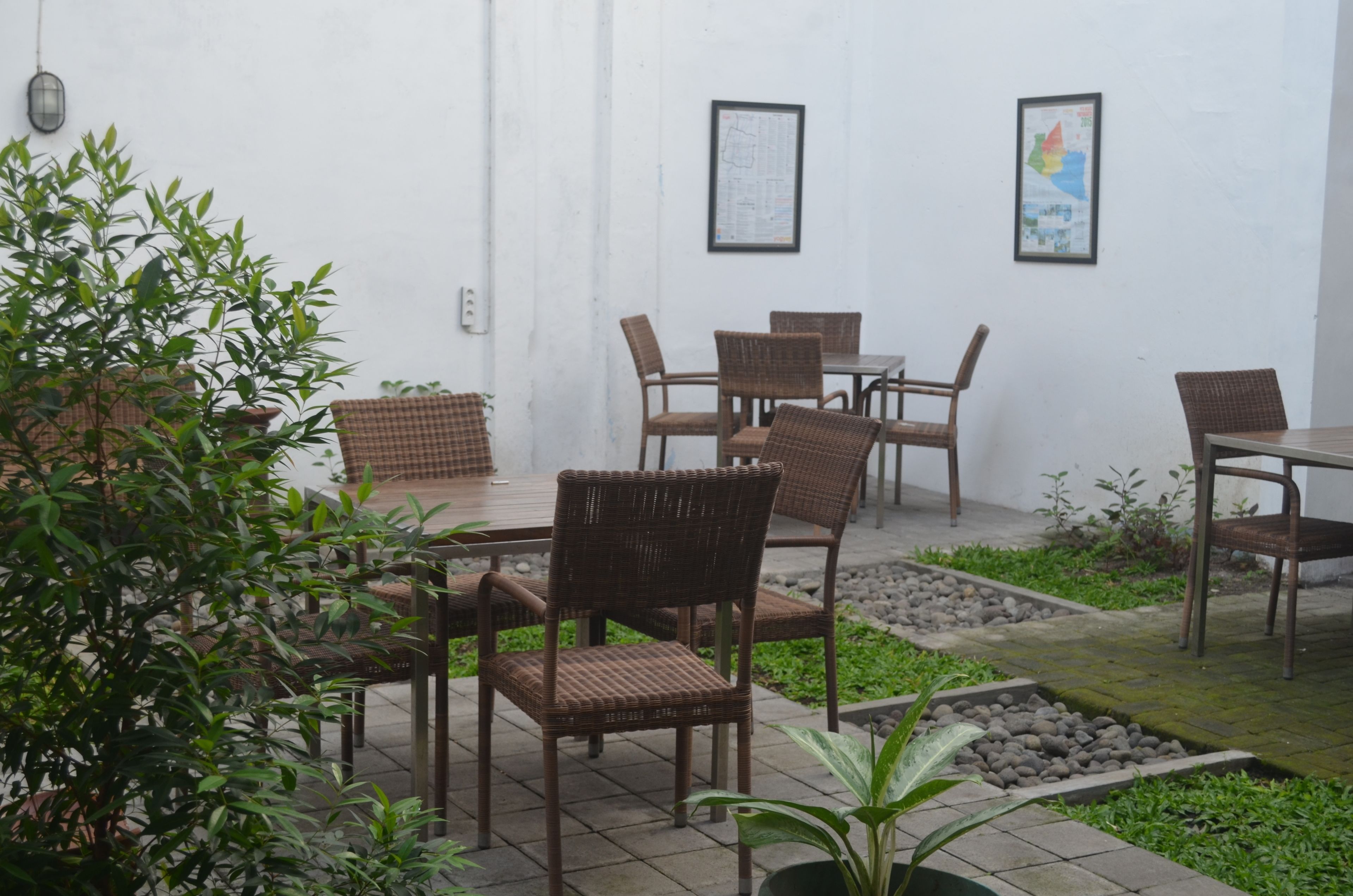 An outdoor seating area with wicker chairs and tables, surrounded by greenery and a white wall with framed artwork.