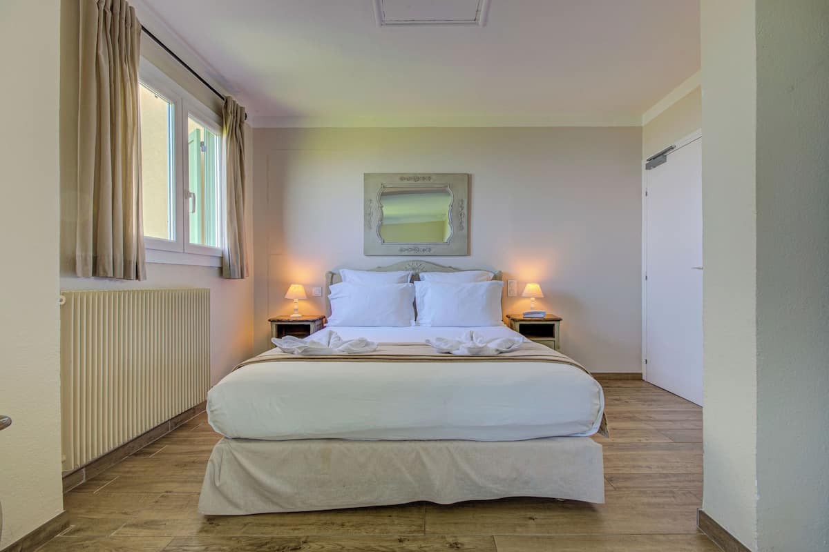 Ein ordentlich bezogenes Bett in einem Hotelzimmer mit Heizkörper, zwei Nachttischlampen und einem gerahmten Spiegel an der Wand.