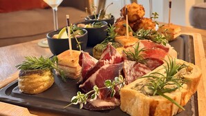 A charcuterie board with various meats, cheeses, and garnishes on a slate tray, accompanied by a glass of white wine and a martini glass. - Hotel Restaurant La Corniche (San-Martino-di-Lota)