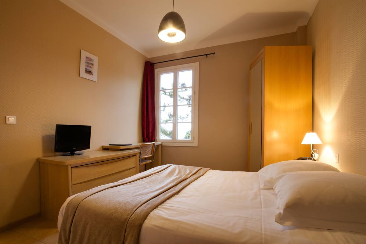 Ein Hotelzimmer mit einem Bett, einem Schreibtisch mit Fernseher, einem Fenster mit Vorhängen und einer Nachttischlampe.