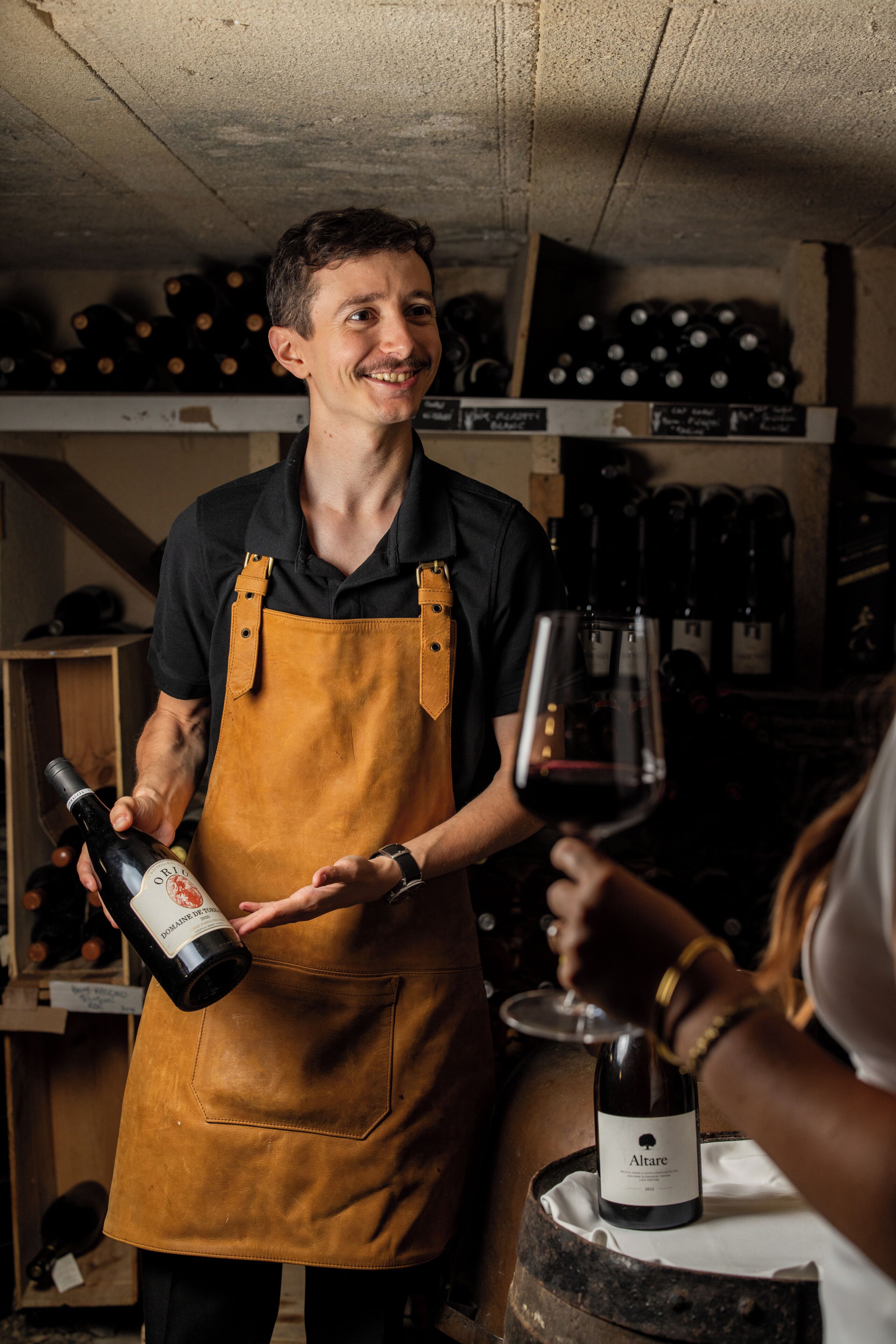 Une personne vêtue d’une chemise noire et d’un tablier marron, tenant une bouteille de vin et un verre, se tient dans une pièce où des bouteilles de vin sont disposées sur des étagères.