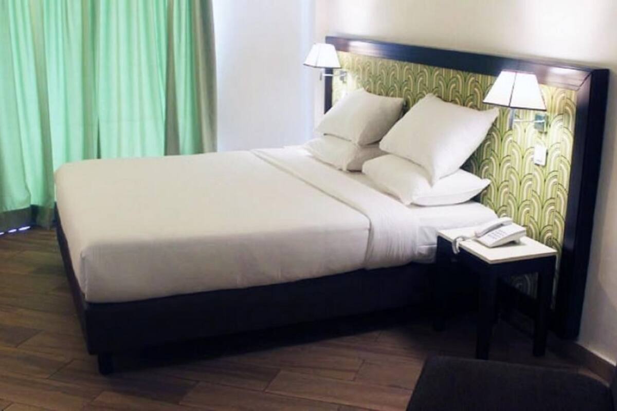 A neatly made bed with white linens and pillows, a bedside table with a lamp, and a patterned wallpaper on the wall.