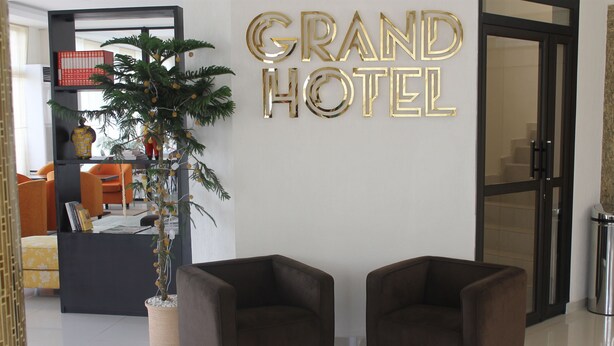 Un vestíbulo con mostrador de recepción, dos sillones marrones, una planta en maceta y un cartel que dice 'Grand Hotel'.
