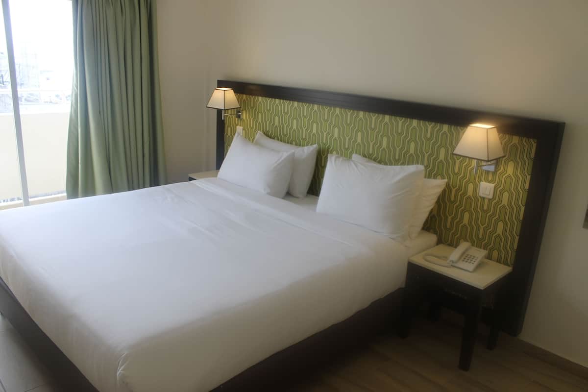 A hotel room with a large bed, two bedside lamps, a bedside phone, and a view of the outdoors through a window with curtains.