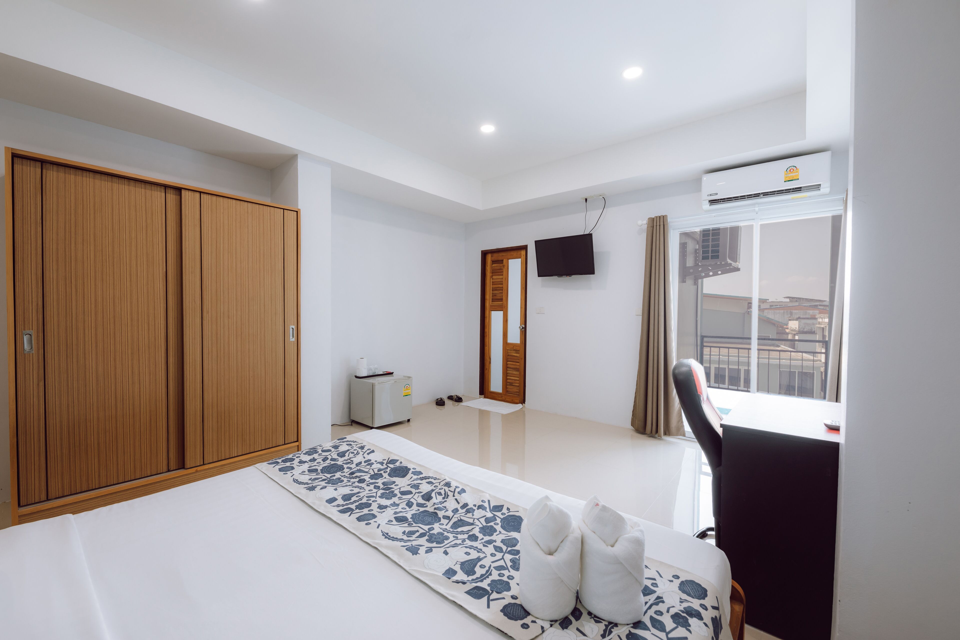 A modern hotel room with a large wardrobe, a flat-screen TV mounted on the wall, a window with a view of the city, and a small desk with a chair.