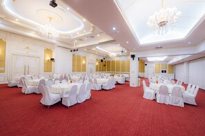 Un salón de banquetes con mesas redondas y sillas blancas, suelo de moqueta roja y detalles ornamentales en el techo.