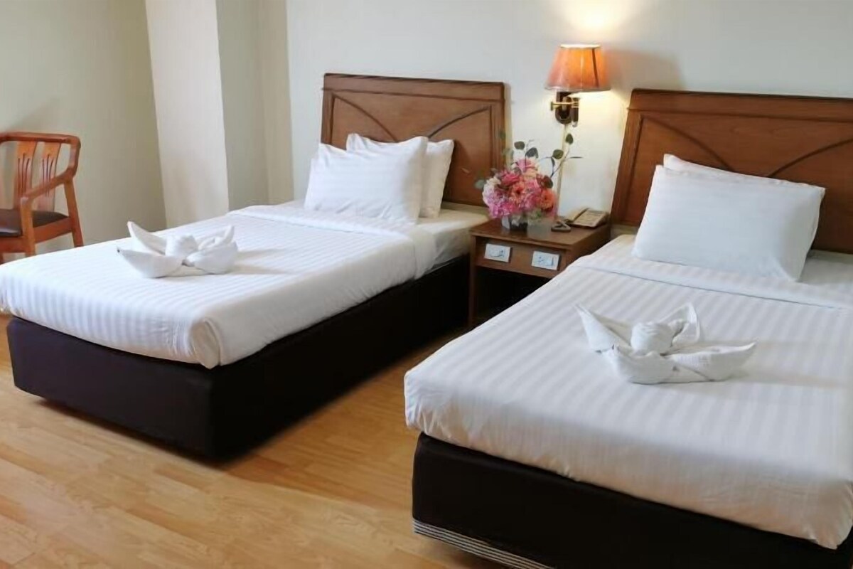 Two single beds with wooden headboards, white linens, and a small table with a lamp and a vase of flowers between them.