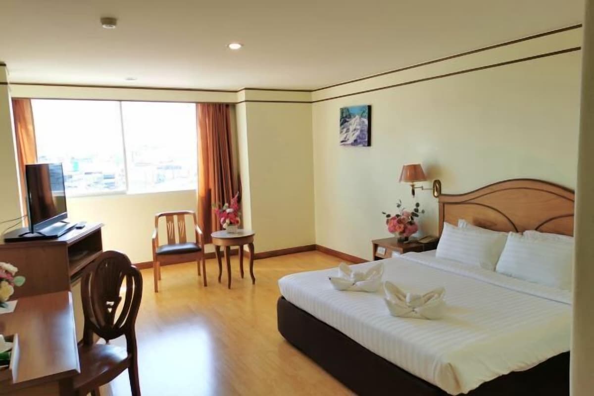 A hotel room with a bed, a desk with a computer, a chair, a small table with flowers, and a window with curtains.