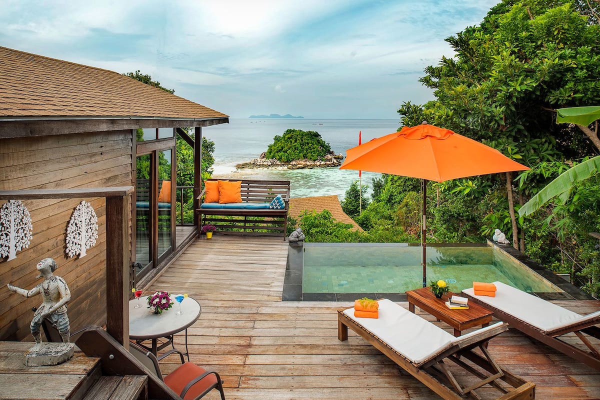 A wooden deck with a glass-bottomed infinity pool, a wooden bench with cushions, and a table with chairs.