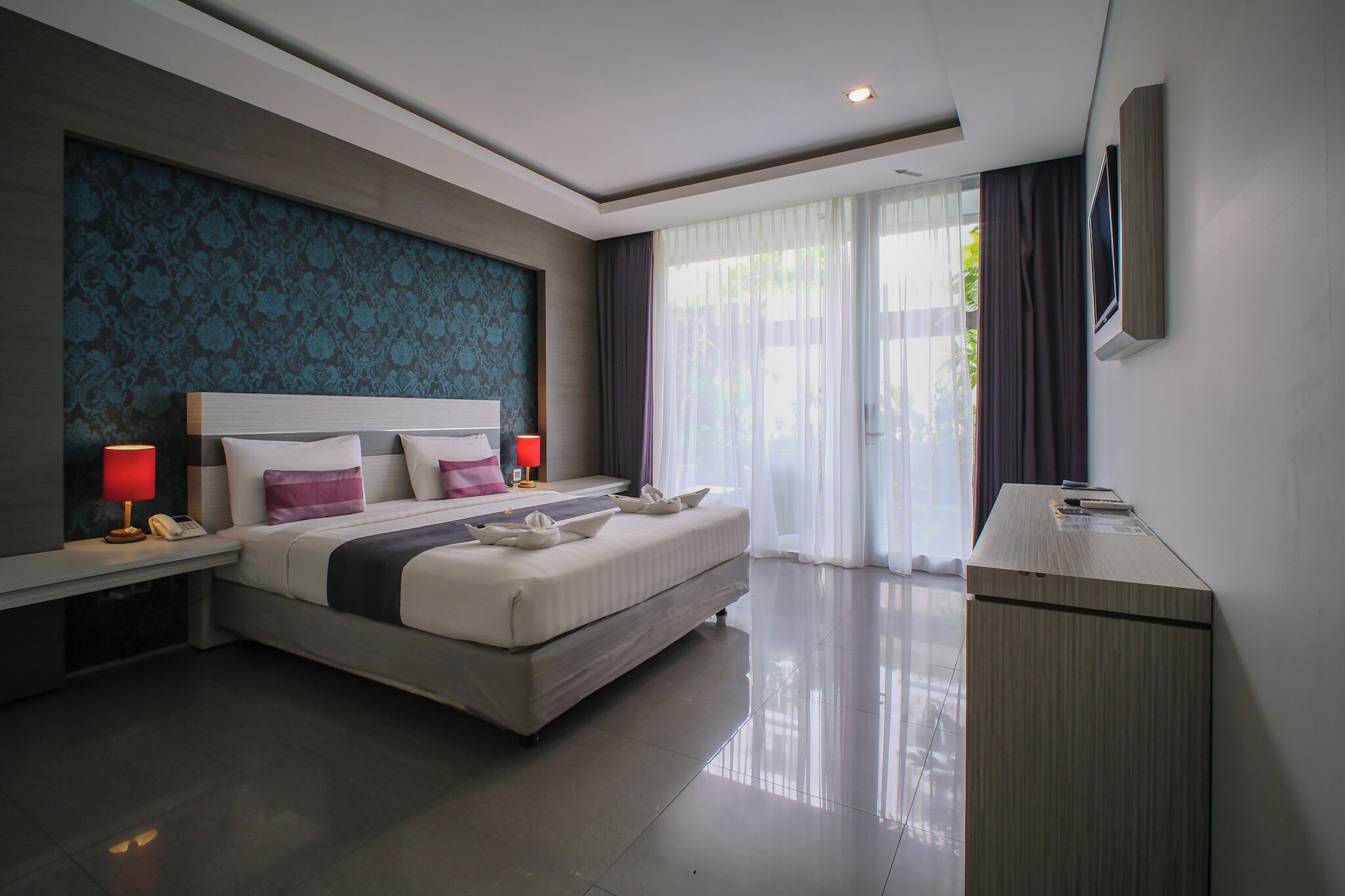 A modern hotel room with a large bed, bedside lamps, a desk, and a view of greenery outside.