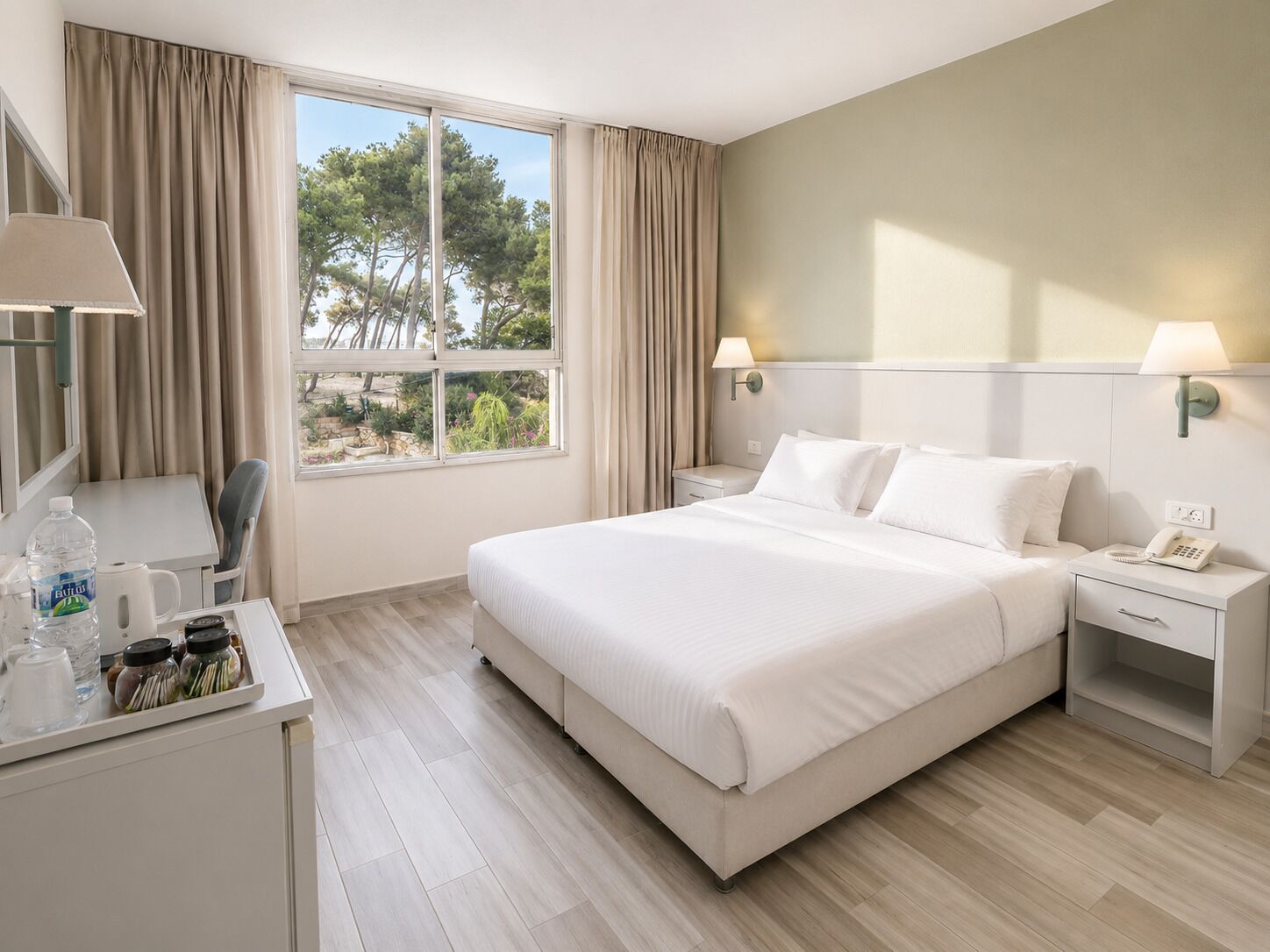 A hotel room with a large bed, bedside tables, a desk with a chair, a window with curtains, and a view of trees.