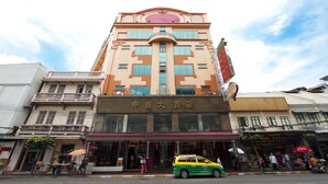 A hotel with a prominent sign in a foreign language, a balcony on the second floor, and a street in front with a yellow taxi. - Chinatown Hostel (Bangkok)