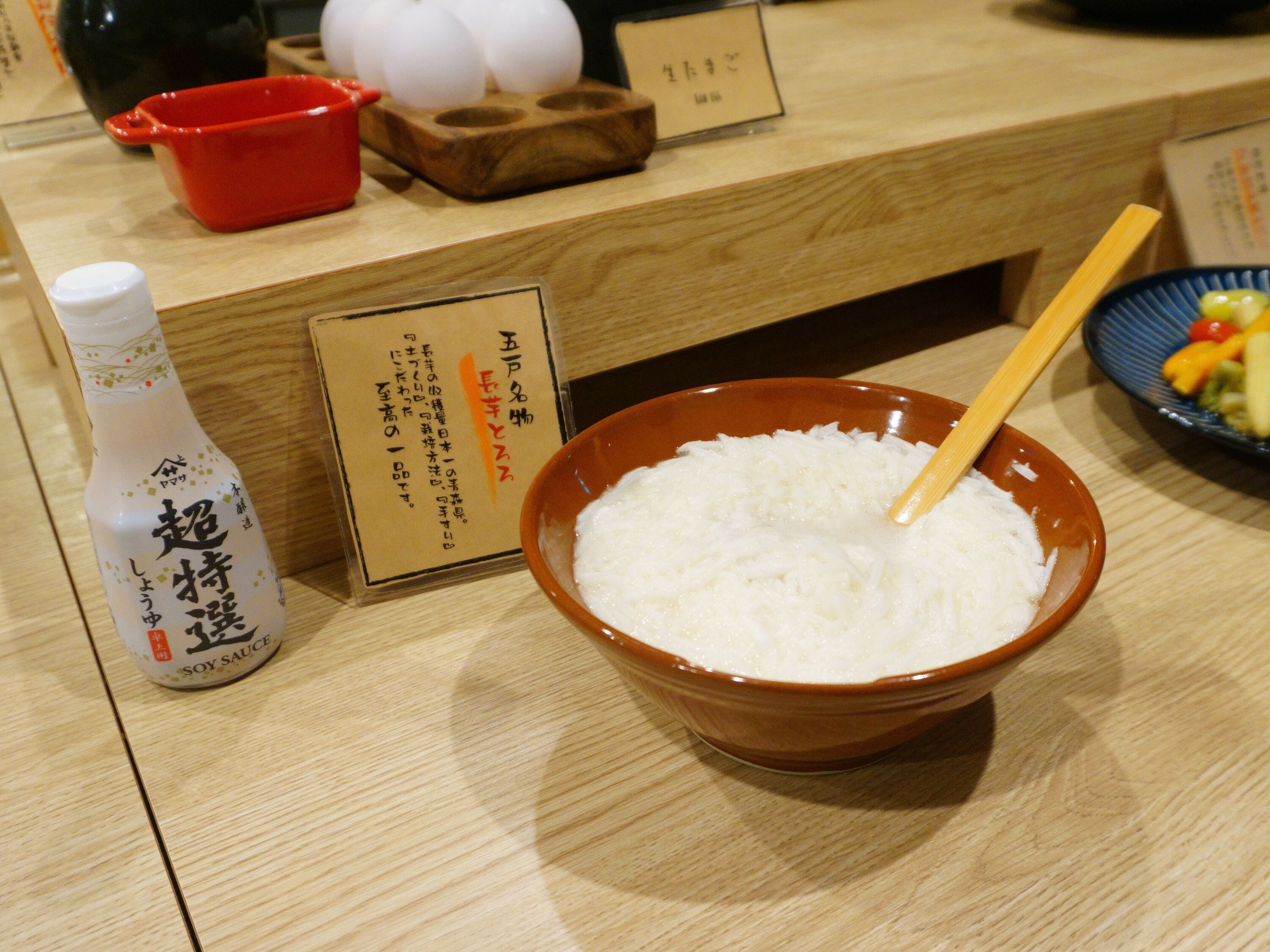 A wooden table with a bowl of white rice, a bottle of soy sauce, and a red container.