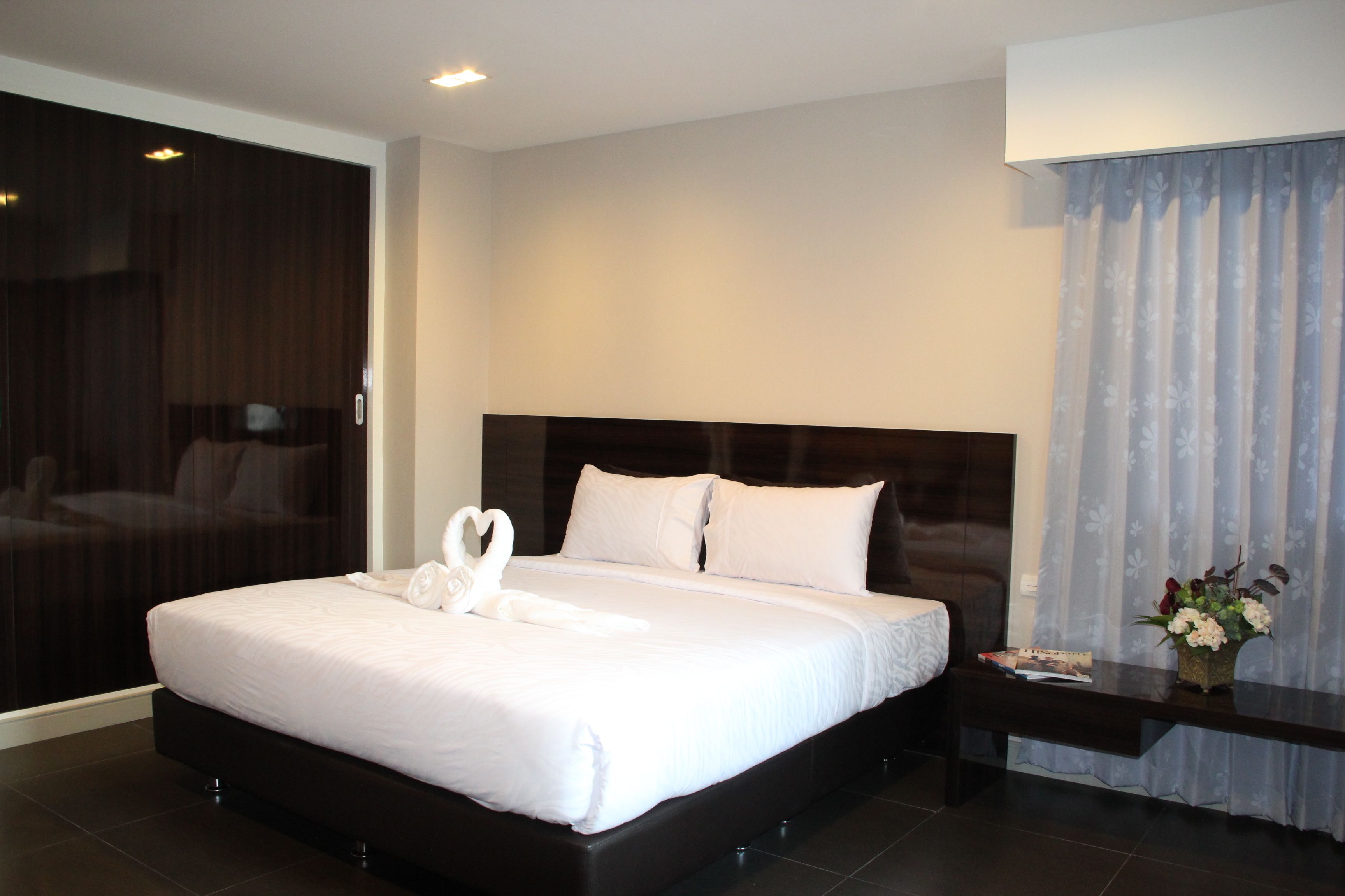 A modern hotel room with a large bed, dark wood headboard, white linens, a nightstand with a vase of flowers, and a large window with sheer curtains.