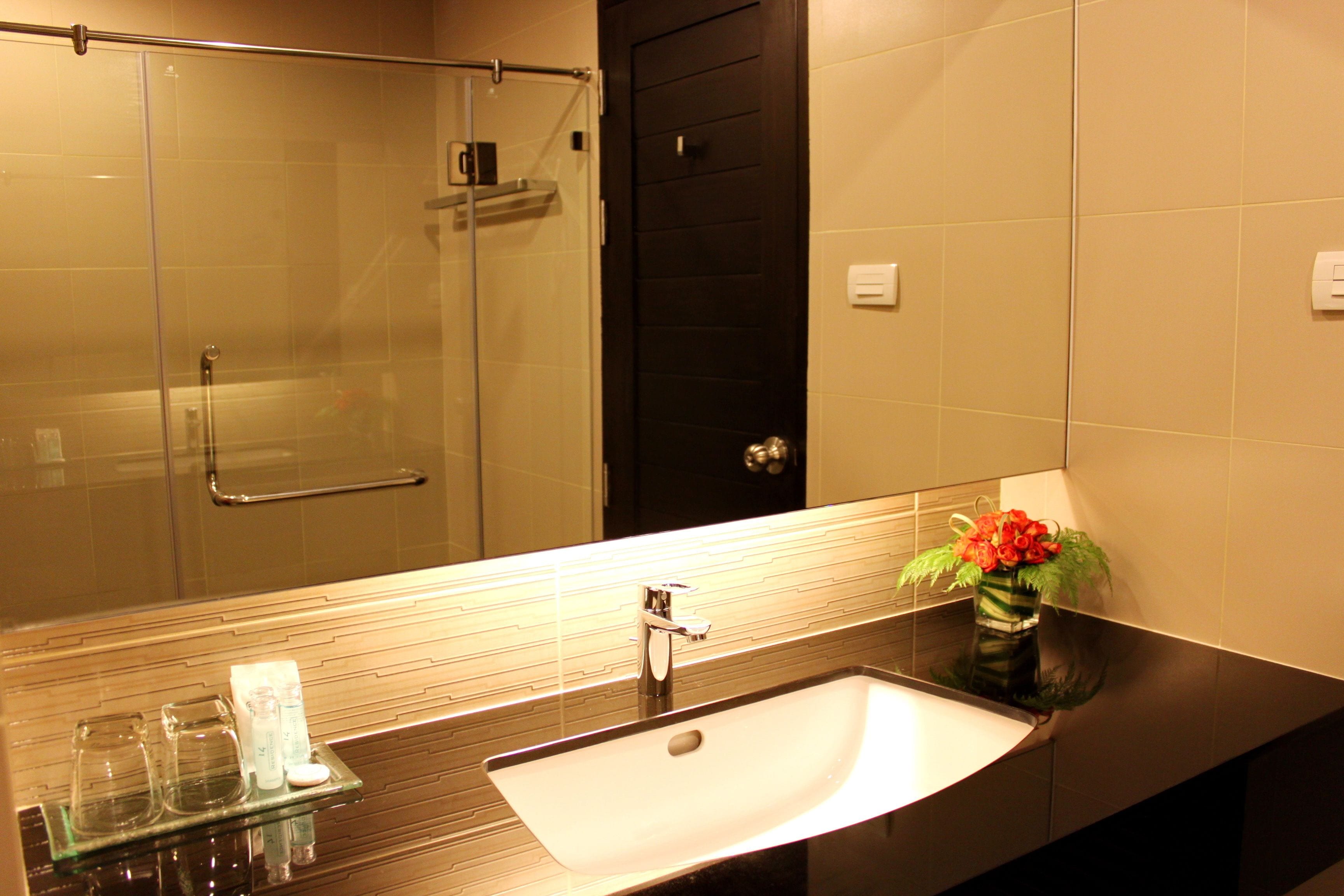 A bathroom with a glass shower enclosure, a black door, a white sink with a glass shelf, and a vase with flowers.