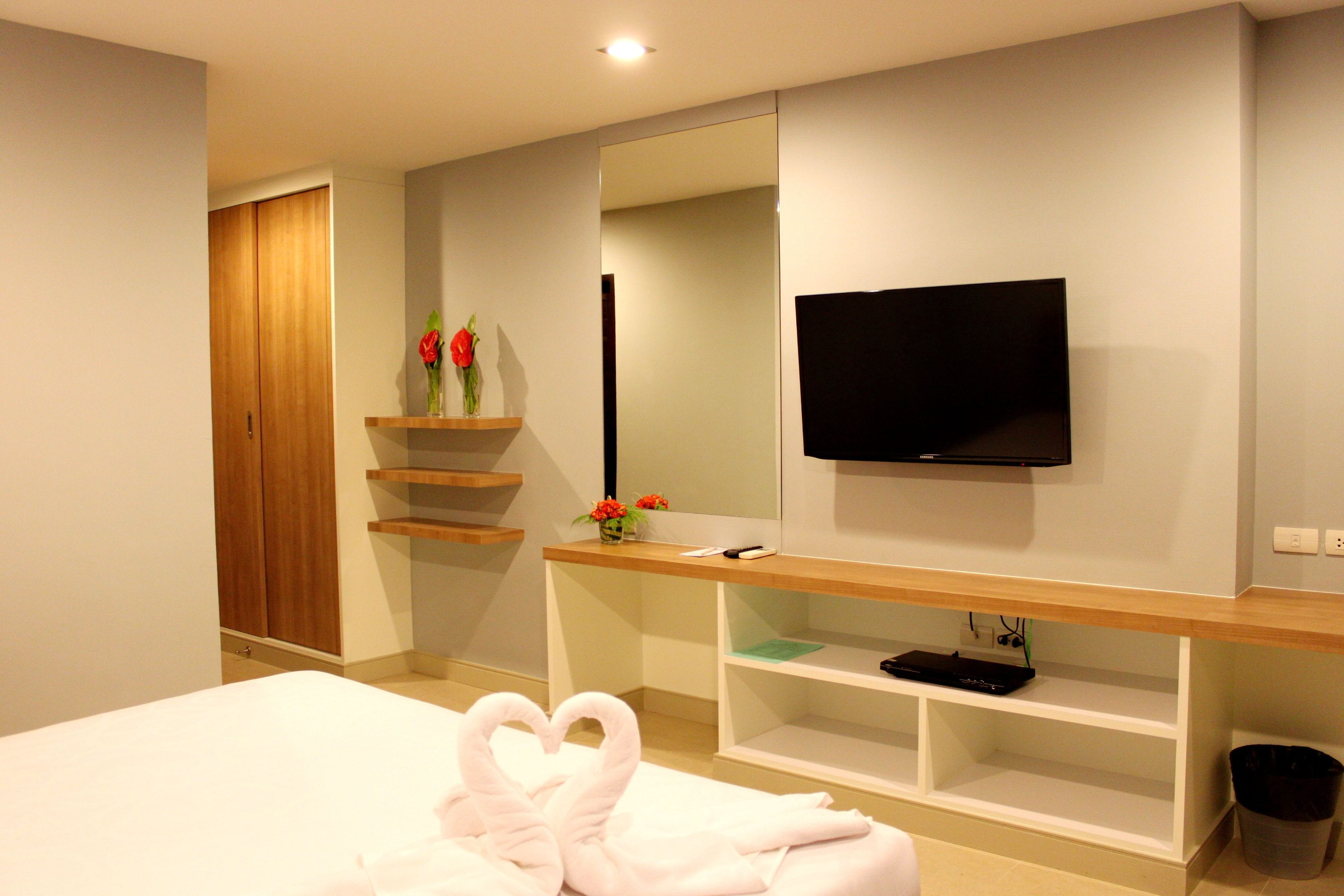 Habitación de hotel con una cama, un televisor montado en la pared, un estante de madera con un florero y un armario de madera.