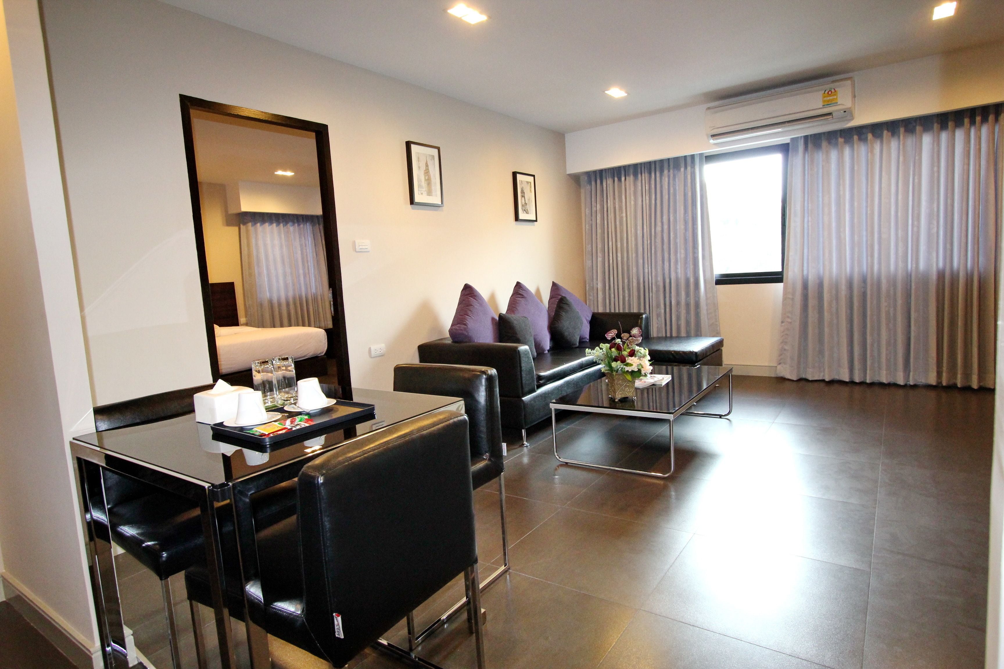Een moderne hotelkamer met een bank, twee fauteuils, een salontafel en een eettafel met stoelen. Er is een grote spiegel, een raam met gordijnen en een airconditioner aan de muur.