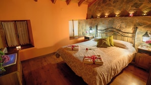A bedroom with a stone wall, wooden ceiling beams, a bed with towels, a nightstand, and a window with curtains. - Entre Valles (Carrocera)