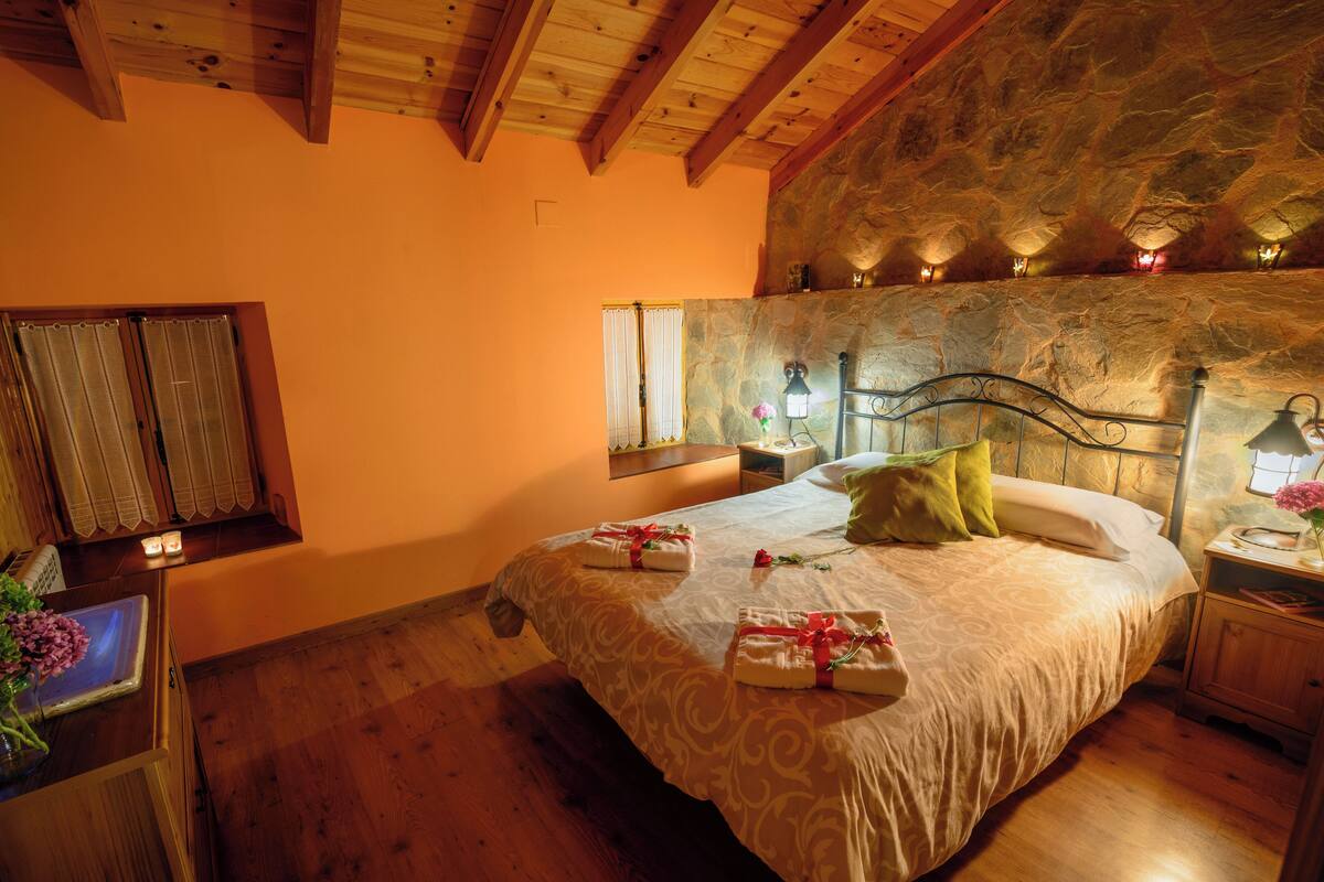 A cozy bedroom with a stone wall, wooden ceiling beams, a large bed with towels and pillows, a nightstand with a lamp, and a window with curtains.