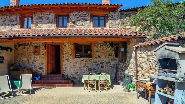 A stone house with a tiled roof, a wooden porch, a dining table with chairs, a barbecue grill, and a small garden area. - Entre Valles (Carrocera)