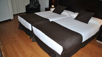 Una cama doble con cabecera oscura y ropa de cama blanca, una mesita de noche con lámpara, y suelo de madera.
