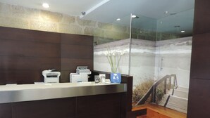 A reception desk with a glass partition, a wooden staircase, and a stone wall backdrop. - Hotel Junquera (Vigo)