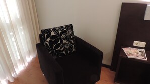 A black armchair with a floral pillow, a wooden floor, a white wall, and a small table with magazines. - Hotel Junquera (Vigo)