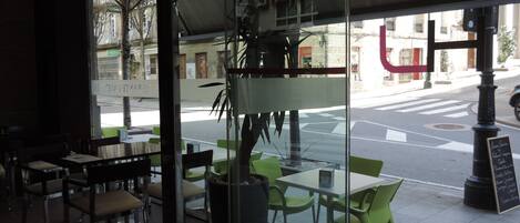 An outdoor seating area with tables and chairs, a large potted plant, and a view of the street.