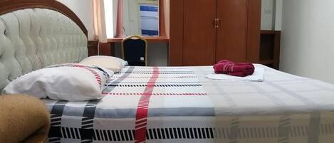 A hotel room with a bed, a wooden headboard, a mirror, a wardrobe, and a desk with a chair.