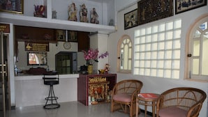 Uma área de recepção com balcão, duas cadeiras de vime, um altar decorativo e uma janela grande com vários painéis.