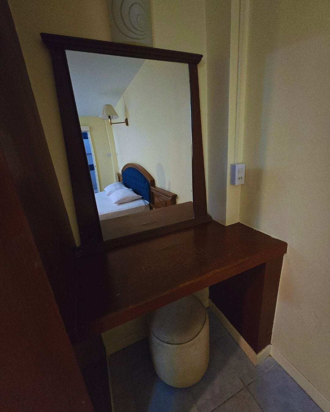 A wooden desk with a mirror, a small stool, and a bed with a blue headboard in the reflection.