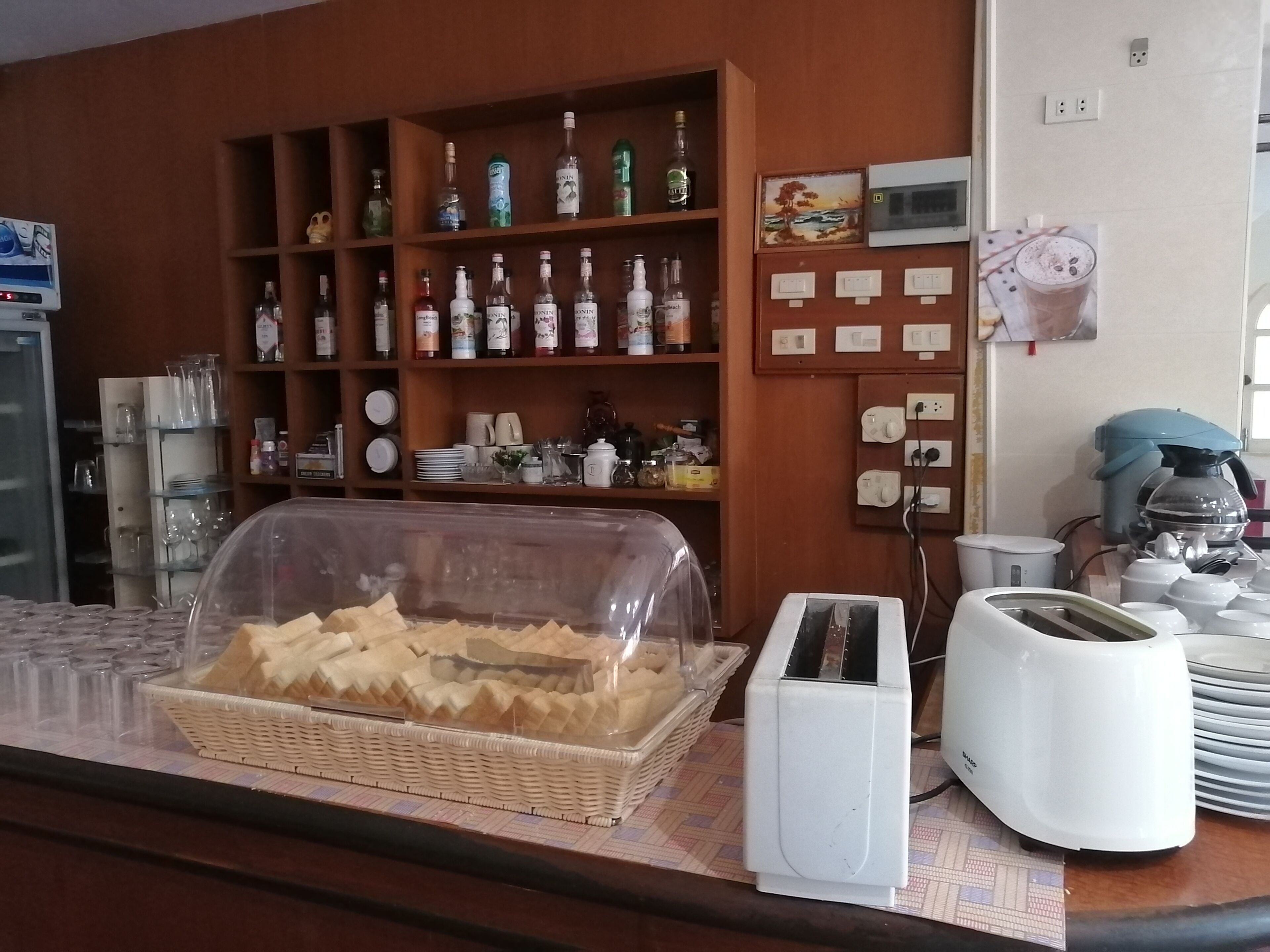 A breakfast bar with a basket of pastries, a toaster, and a coffee machine. Behind the bar, there are shelves with various bottles and kitchenware.