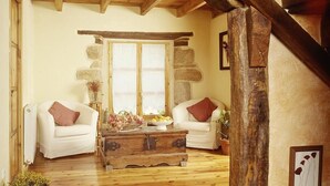 Una habitación acogedora con suelo de madera, dos sofás blancos con cojines marrones, una mesa de centro de madera rústica y un amplio ventanal con pilares de piedra.
