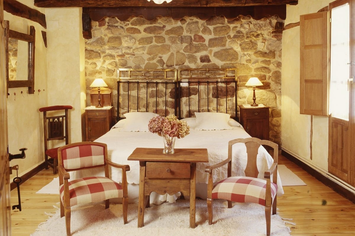 Une chambre à coucher au style rustique, avec un mur en pierre, des poutres en bois au plafond, un grand lit avec deux tables de chevet, deux chaises et une petite table sur laquelle est posé un vase de fleurs.
