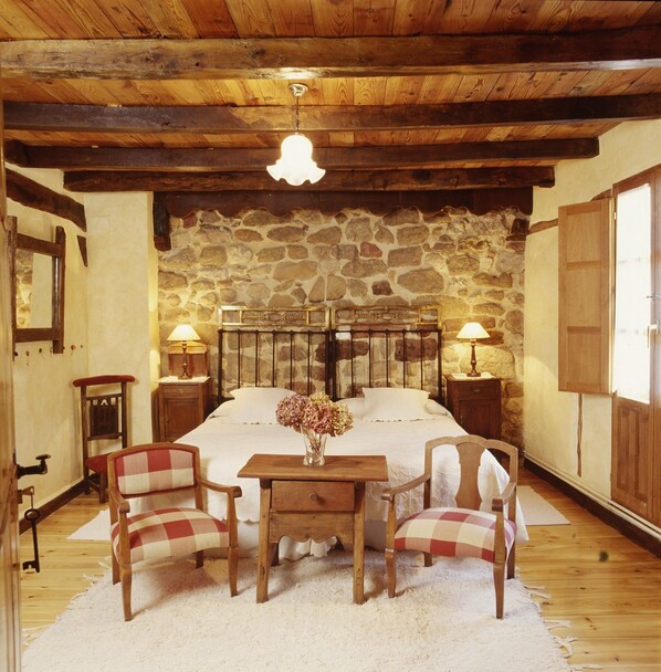A rustic bedroom with a stone wall, wooden ceiling beams, a large bed with two bedside tables, two chairs, and a small table with a vase of flowers.
