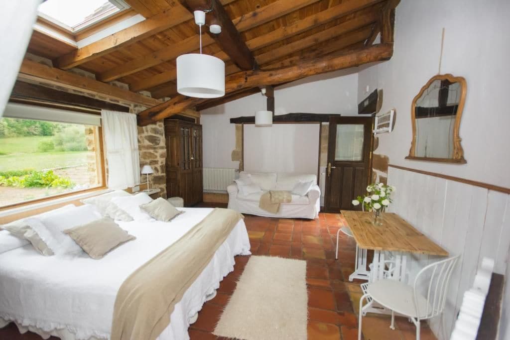 A bedroom with a large bed, a wooden ceiling, a stone wall, a white sofa, and a small table with a vase of flowers.